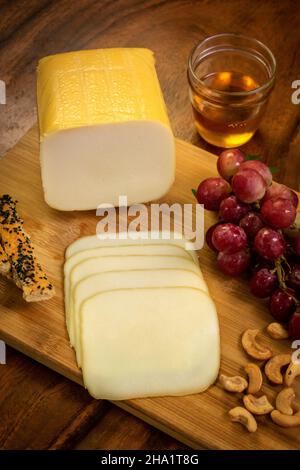 Geräucherter Mozzarella-Käseblock auf Holzbrett mit Trauben, Cashewnüssen, Brotscheiben und Honig Stockfoto
