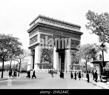 Paris Arc de triomphe 1945 mit Fußgängern, Zylisten und wenigen Autos der Blick auf den Bogen und den kreisförmigen Place de l'Étoile ist von der Avenue Marceau gegen Ende des Zweiten Weltkriegs in Europa aufgenommen. Auf der Fahrbahn befinden sich nur mehrere Kraftfahrzeuge und mehrere Radfahrer. Auf dem Foto sind zahlreiche Fußgänger im Vordergrund und Besucher unter dem Arc zu sehen. Es gibt zwei Kiosk, an denen die Loterie Nationale gefördert wird. Alte Straßenlaternen sind zu sehen. Das Foto wird von Bäumen mit Frühlingslaub eingerahmt. Stockfoto