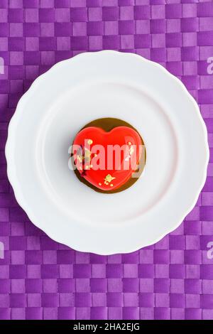 Ein Kuchen in Herzform. Ein herzförmiger Mousse-Cupcake auf einem Teller auf violettem Hintergrund. Valentinstag. Vertikales Foto Stockfoto