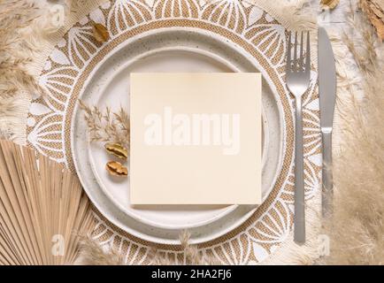 Hochzeitstisch Platz mit quadratischer unbeschriftete Karte auf Platte auf böhmischen Tischset Draufsicht. Boho Hochzeitskarte Mockup. Tropisch-beige Schablone mit getrockneten Pflanzen Stockfoto