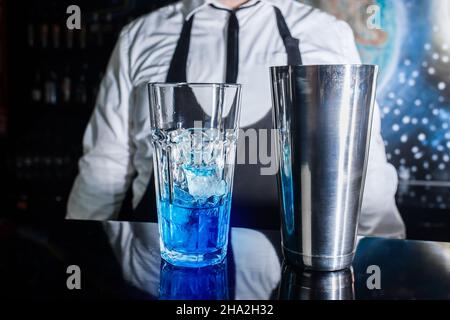 Ein Glas mit Eis und blauem Sirup steht auf der Theke vor dem Hintergrund eines professionellen Barkeepers, neben einem Werkzeug zum Mischen und Zubereiten Stockfoto