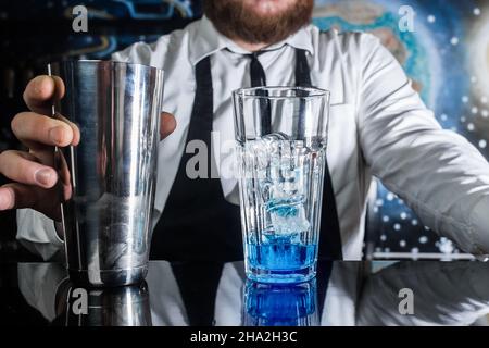 Der Prozess der Zubereitung eines Cocktails. Die Hand eines professionellen Barkeepers hält ein flüssiges Metallschüttler-Mischwerkzeug neben einem Glas mit Eis und blauem sy Stockfoto