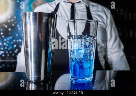 Ein Glas mit Eis und blauem Sirup steht auf der Theke vor dem Hintergrund eines professionellen Barkeepers, neben einem Werkzeug zum Mischen und Zubereiten Stockfoto