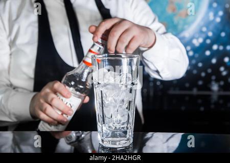 Der Prozess der Zubereitung eines Cocktails. Professioneller Barkeeper mit einer Flasche Sirup in den Händen neben einem Glas Eiswürfel auf der Theke. Stockfoto