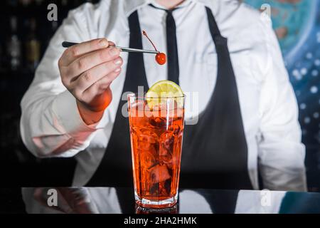 Ein professioneller Barkeeper steckt eine Kirsche in einen roten gekühlten alkoholischen Cocktail mit einer Barpinzette im Glas an einer Nachtclub-Theke. Stockfoto