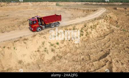 Muldenkipper fährt auf Landstraße. Szene. Draufsicht auf LKW-Fahrten, die vor dem Hintergrund der Baggerkarriere Staubwolken auf unbefestigten Straßen auf dem Land hinterlassen Stockfoto