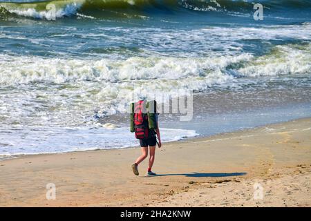 Junge Backpacker-Frau, die auf dem Meer unterwegs ist. Mädchen Reisende mit großen roten Rucksack erkunden neue Region, schöne Ostsee Hintergrund. Solo weiblich t Stockfoto