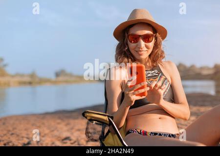 Glückliche Frau kommuniziert mit einem Freund auf einem Smartphone, während sie auf dem Campingstuhl am Flussufer sitzt. Sommerferien am Wasser und mobile Inter Stockfoto