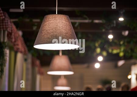 Pendelleuchten über Tischen im Café der Stadt, Restaurant mit Abendbeleuchtung. Beigefarbene Lampenschirme aus Stoff mit schwach gedimmtem warmem Licht. Moderne gemütliche Innenausstattung, Land Stockfoto