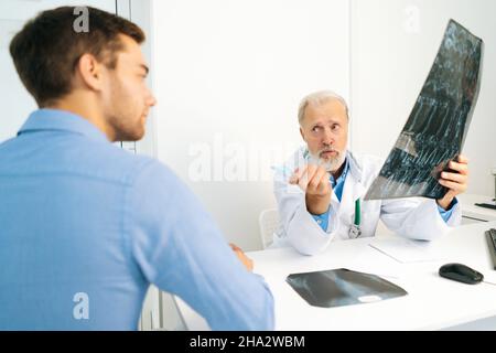 Blick vom Rücken eines jungen männlichen Patienten, der von einem professionellen, älteren dult-Arzt hört, der die Ergebnisse der MRT-Untersuchung erklärt Stockfoto