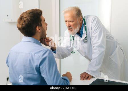 Ansicht von hinten zu reifen erwachsenen männlichen Arzt otolaryngologist Durchführung einer Untersuchung des jungen Mannes Patienten Hals im Büro der medizinischen Klinik. Stockfoto