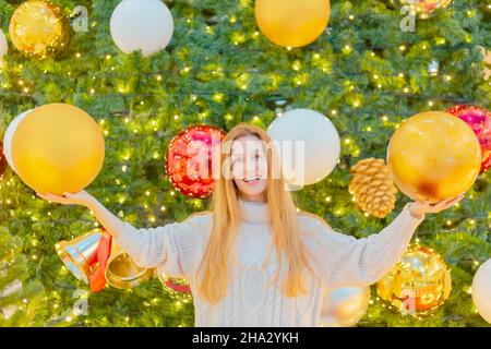 Eine fröhliche Dame hält riesige Weihnachtskugeln vor dem Hintergrund eines eleganten glitzernden Weihnachtsbaums. Festliches Poster. Herzlichen Glückwunsch Stockfoto