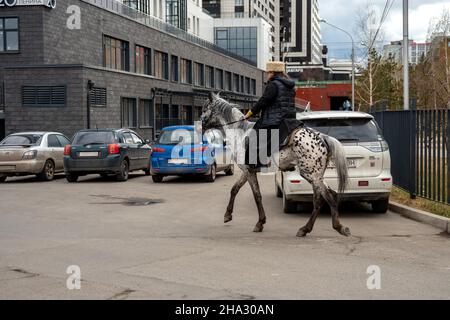 Ein Geistlicher in einer Soutane und einem Pelzhut reitet auf einem Pferd an geparkten Autos entlang einer städtischen Frühlingsstraße vorbei. Stockfoto