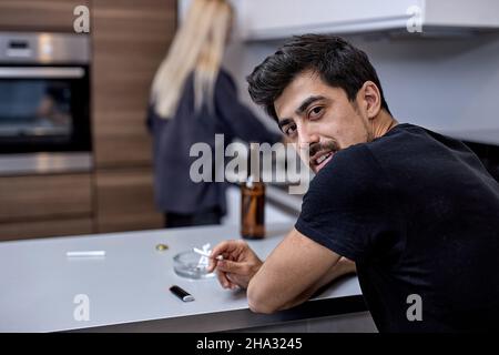 Paar mit Problemen mit Meinungsverschiedenheiten in der Küche, Mann sitzen Rauchen und Bier Alkohol trinken, während Frau kocht. Konzentrieren Sie sich auf Brunette Bartmann. Stockfoto