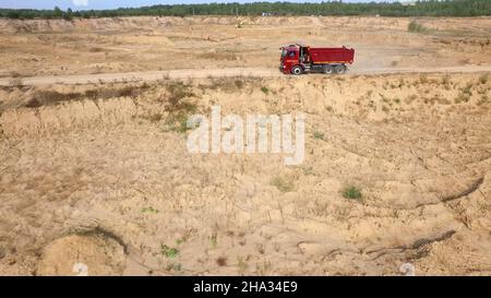 Muldenkipper fährt auf Landstraße. Szene. Draufsicht auf LKW-Fahrten, die vor dem Hintergrund der Baggerkarriere Staubwolken auf unbefestigten Straßen auf dem Land hinterlassen Stockfoto