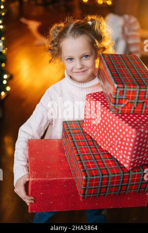 Vertikales Porträt von fröhlichen kleinen blonden Mädchen hält viele Geschenkboxen mit Weihnachtsgeschenken in gemütlichen Wohnzimmer mit festlichen Innenraum. Stockfoto