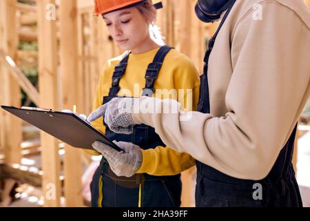 Architektin und Zimmermann diskutieren die Baupläne, während sie in einem unfertigen Haus stehen, Architektin und Baumeister, die in einer Arbeitsuni gekleidet sind Stockfoto