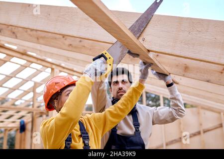 kaukasische selbstbewusste weibliche Auftragnehmerin in orange Hardhat mit Säge Hand schneiden Holzbalken in Holzhaus Website. Junger, ansprechender bärtiger Mann, Baumeister Stockfoto