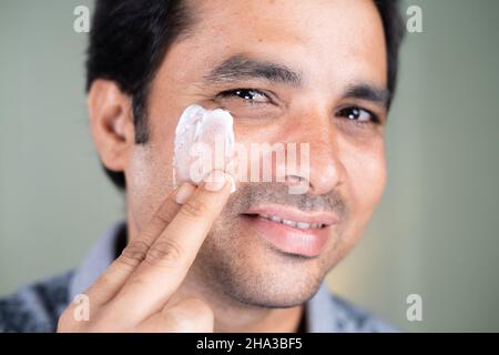 Nahaufnahme des gut lächelnden jungen Mannes, der zu Hause Creme oder Anti-Aging-Feuchtigkeit auf das Gesicht aufgibt, indem er die Kamera anschaut - Konzept der Fairness und Stockfoto