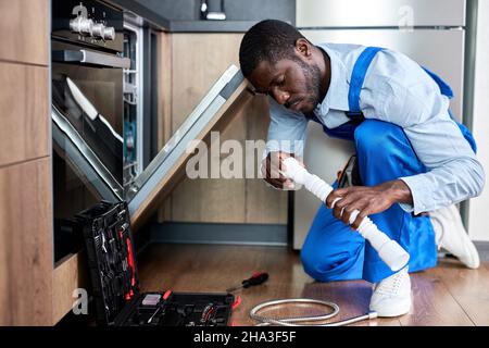 Seitenansicht Young African Handyman Repariert Den Geschirrspüler, Wechselt Den Siphon Und Trägt Blaue Arbeitskleidung. Selbstbewusster Professioneller Handyman Ar Home, Fokus Stockfoto