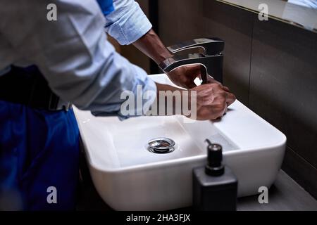 Nahaufnahme der Hand eines afroamerikanischen schwarzen Klempners, der versucht, einen Wasserhahn im Badezimmer zu öffnen, repariert ein professioneller Handwerker oder Klempner in blauer Uniform i Stockfoto