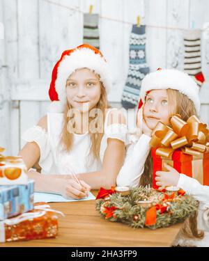 Zwei kleine Mädchen Schwestern in weihnachtsmännern sitzen am Tisch und schreiben einen Brief an den weihnachtsmann, träumen von Geschenken in der weihnachtsnacht, schaffen eine Wunschliste, der Tisch ist mit .Weihnachtskranz, Kerzen und voller Weihnachtsgeschenke geschmückt. Weiße Holzwand mit Weihnachtsstrumpf auf dem Hintergrund. Nahaufnahme . Hochwertige Fotos Stockfoto