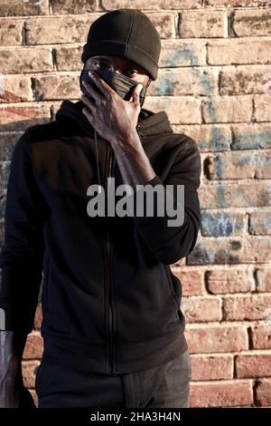 Porträt eines jungen afrikanischen Mannes in Maske stehen an der Ziegelwand, Hooligan in schwarzer Kleidung. Schwarzer Kerl stellen eine Gefahr für die Gesellschaft, warten auf Opfer, begehen Stockfoto