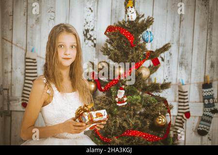 kaukasische Mädchen-Modell mit langen welligen blonden Haaren in einem weißen flauschigen Kleid mit einem Geschenk in den Händen steht neben einem Weihnachtsbaum mit Kugeln verziert. Mädchen schaut auf die Kamera im Weihnachts-Neujahrsstudio. Im Hintergrund sind Weihnachtssocken mit Ornamenten an der Wand zu sehen. Nahaufnahme. Hochwertige Fotos Stockfoto