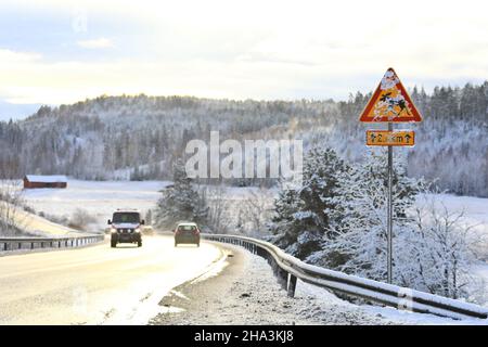 Elchwarnschild teilweise mit Schnee bedeckt mit einem Krankenwagen, der auf nasser Straße durch die winterliche Landschaft in Finnland fährt. Stockfoto