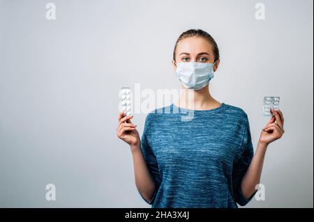 Junge Frau in einer medizinischen Maske auf weißem Hintergrund. Hält eine Blister von Pillen aus dem Virus. Blick direkt auf die Kamera. Epidemie, Coronavirus, Stockfoto