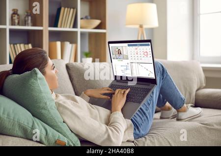 Frau, die zu Hause auf dem Sofa liegt, am Laptop arbeitet, den Kalender nutzt, im Arbeitsgespräch kommuniziert Stockfoto