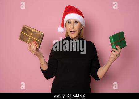 Ältere Frau trägt weihnachtsmann Hut hält Geschenkboxen isoliert auf rosa Hintergrund. Stockfoto