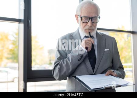 Ein nachdenklicher Geschäftsmann hält den Stift, während er den Vertrag im Büro liest Stockfoto