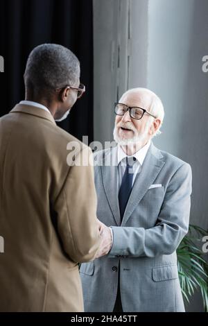 Positiver Senior Businessman in Brillen, die sich mit einem afroamerikanischen Kollegen im Büro die Hände schütteln Stockfoto