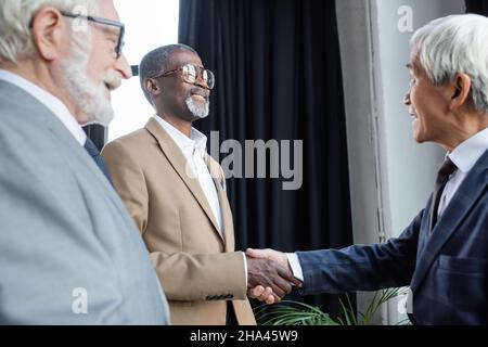 Verschwommener Senior-Geschäftsmann lächelt in der Nähe afroamerikanischer und asiatischer Geschäftspartner, die sich im Büro die Hände schütteln Stockfoto