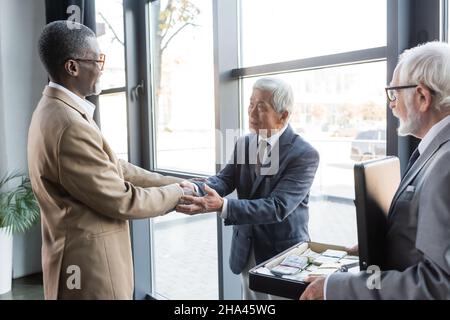 Älterer Geschäftsmann hält eine Aktentasche mit Geld, während interrassische Geschäftspartner im Büro die Hände schütteln Stockfoto