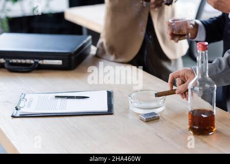 Beschnittene Ansicht von verschwommenen Geschäftsleuten mit Zigarre und Whiskey in der Nähe von Vertrag und Aktentasche auf dem Schreibtisch Stockfoto