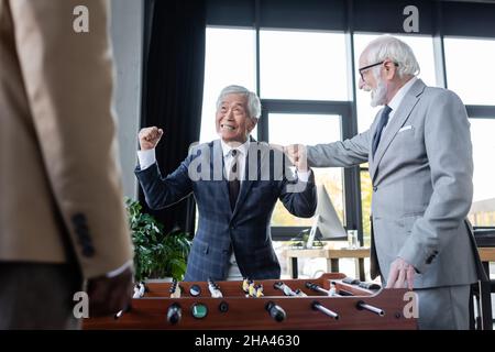 Glücklicher asiatischer Geschäftsmann, der in der Nähe des Tischfußballs und hochrangiger multiethnischer Kollegen eine Siegesture zeigt Stockfoto