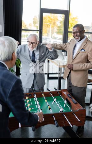 Aufgeregter Geschäftsmann, der in der Nähe von Tischfußball und multikulturellen Kollegen eine Triumphgeste zeigt Stockfoto