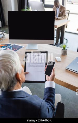 High-Angle-Ansicht des Geschäftsmannes, der mit Vertrag arbeitet, während er das Smartphone mit leerem Bildschirm hält Stockfoto