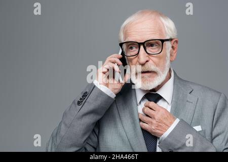 Ein erfahrener, nachdenklicher Geschäftsmann rührt Krawatte an, während er auf einem Mobiltelefon auf grauem Telefon spricht Stockfoto