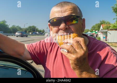 Portarait von hungrigen kaukasischen Senior-Fahrer in Sonnenbrillen essen Junk-Food (gebratenes Patty) in der Nähe seines Autos, während der kurzen Halt auf der ukrainischen Straße Stockfoto