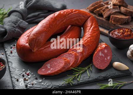 Geräucherte Wurst mit geschnittenen Stücken auf einem Schieferbrett mit Rosmarin, Knoblauch und Gewürzen. Seitenansicht, Nahaufnahme. Stockfoto