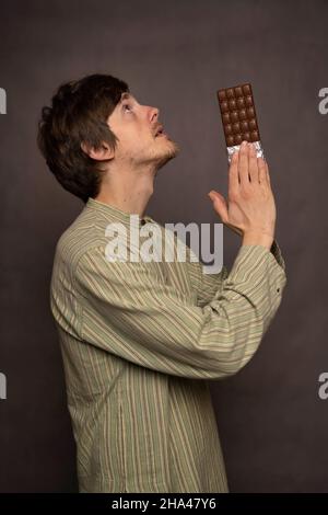 Junger, hübscher, großer, schlanker weißer Mann mit braunem Haar, der mit einem Schokoriegel in den Händen im gestreiften Hemd auf grauem Hintergrund betet Stockfoto