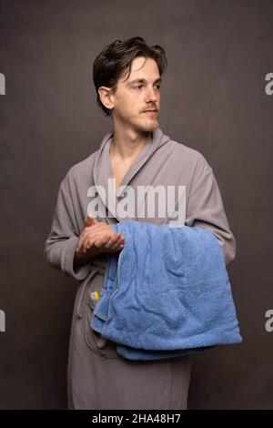 Junger, hübscher, großer, schlanker, weißer Mann mit braunen Haaren, das Handtuch in einem grauen Bademantel auf grauem Hintergrund hält Stockfoto