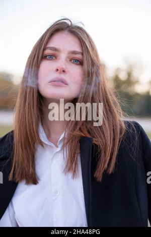 Porträt einer langhaarigen weißen europäerin im dunklen Mantel im Herbst am Strand mit Zigarettenrauch davor Stockfoto