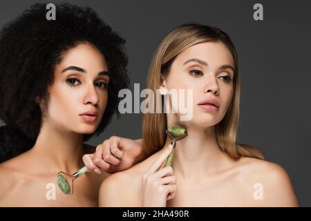 Junge Frau massiert Hals mit Jade Roller in der Nähe afroamerikanischen Freund mit perfekter Haut isoliert auf grau Stockfoto