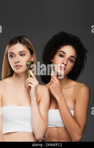 Junge multiethnische Frauen massieren Gesichter mit Jadewalzen, die auf Grau isoliert sind Stockfoto