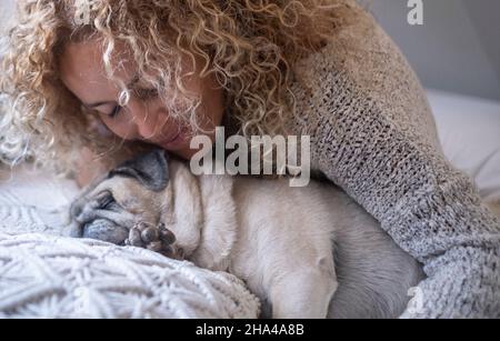 Junge Frau umarmt ihren Hund, während sie auf dem Bett schläft. Nahaufnahme von Frau und ihrem Hund Pug schlafen auf dem Bett zusammen. Frau und Hund schlafen auf gemütlichen Bett zu Hause Stockfoto