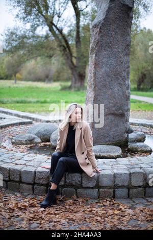 Weiße junge europäische Frau mit blonden Haaren im Park, die auf einem Steinzaun in beigem Herbstmantel sitzt Stockfoto